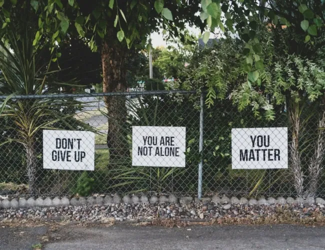 Three signs on a chain-link fence among the trees display heartfelt messages: "Don't Give Up," "You Are Not Alone," and "You Matter." These white signs with black text provide hope and support, essential reminders for anyone managing depression.