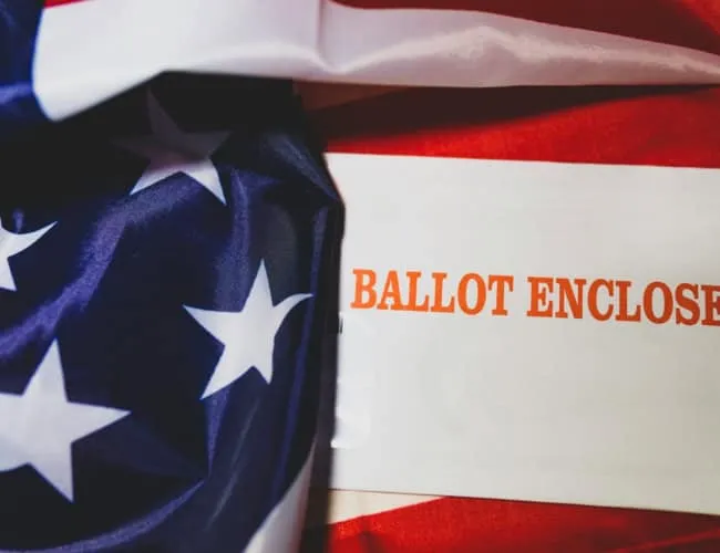 An American Flag with an envelope that says "ballot enclosed" sitting on top of it.