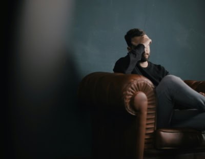 A man dressed in dark jeans and a dark shirt is sitting on a couch with his hand over his eyes.